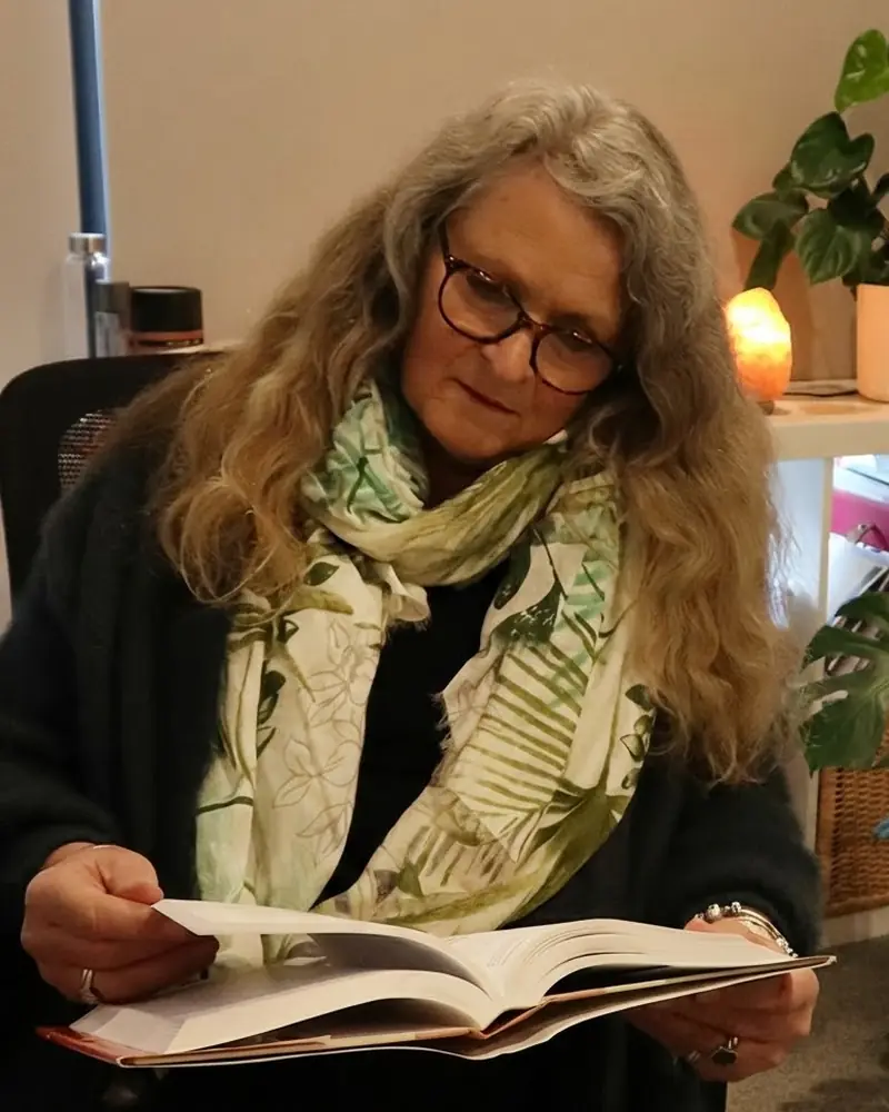Cheryl reading a book in her practice room, beside a salt lamp and indoor plants